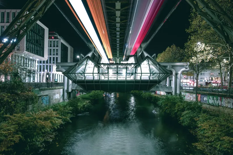 Atmosphärische Nachtaufnahme der Wuppertaler Schwebebahn über der Wupper. Moderne Stationsarchitektur mit illuminierter Glaskonstruktion, dynamischen Lichtstreifen und urbaner Skyline spiegelt sich im Fluss. Einzigartiges Wahrzeichen der Stadt in faszinierender Abendstimmung.
