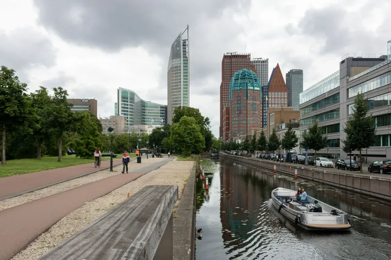 Urbane Stadtansicht von Den Haag mit imposanter Skyline, charakteristischen Hochhäusern und typisch niederländischer Wasserstraße. Das Bild zeigt die perfekte Verschmelzung von moderner Architektur, nachhaltiger Mobilität mit Fahrradwegen und städtischer Natur entlang des Kanals - ein authentischer Einblick in das Leben der niederländischen Regierungsstadt.
