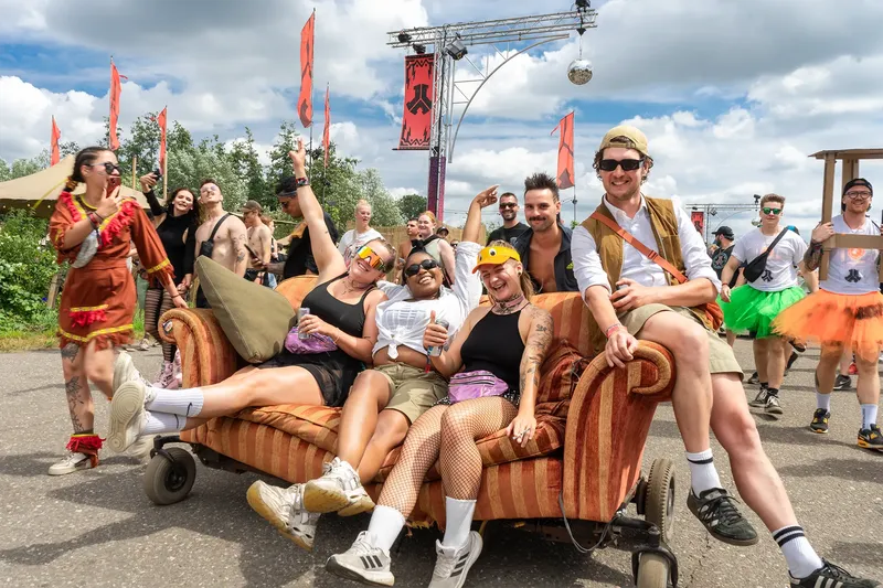 Ausgelassene Festival-Stimmung beim Defekt Festival: Festivalbesucher feiern auf einem rollenden Vintage-Sofa, umgeben von charakteristischen orangenen Fahnen und Festival-Dekoration. Authentische Festivalatmosphäre mit kreativen Outfits und fröhlicher Atmosphäre unter sommerlichem Himmel.
