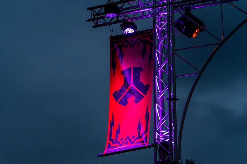 Defqon1 Event-Banner mit futuristischem Logo auf rot-violettem Hintergrund, umrahmt von geometrischen Blitz-Elementen und professioneller Bühnentechnik. Atmosphärische Nachtaufnahme mit LED-Beleuchtung in Violett-Tönen. Ideale Visualisierung für Veranstaltungstechnik und Event-Marketing.
