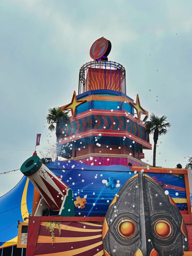 Bunte Festivalbühne mit künstlerischer Dekoration, großer Kanone und stilisiertem Maskengesicht, palmengesäumte Open-Air-Veranstaltung unter bewölktem Himmel, Intents Festival 2025
