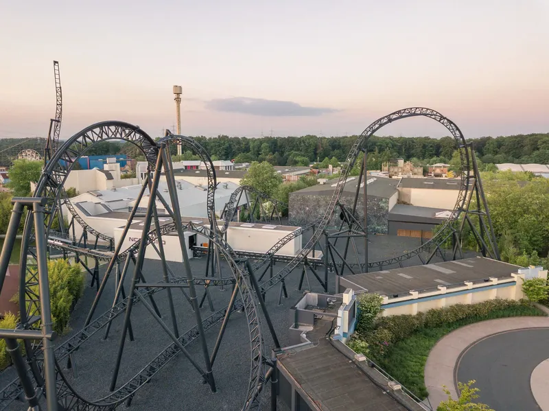 Spektakuläre Luftaufnahme einer modernen Stahl-Achterbahn mit imposanten Loopings und architektonischen Bögen in einem Freizeitpark bei Sonnenuntergang. Die dynamische Stahlkonstruktion präsentiert sich vor einem malerischen Abendhimmel inmitten einer grünen Parklandschaft.
