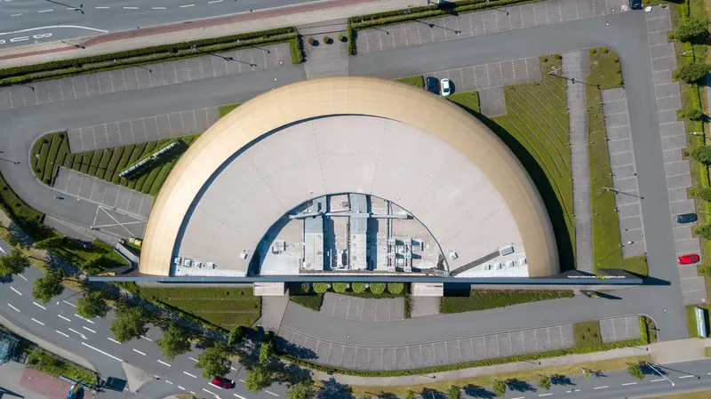 Spektakuläre Drohnenaufnahme einer modernen Kuppelarchitektur aus der Vogelperspektive. Das eindrucksvolle Bauwerk präsentiert sich mit einer markanten halbrunden Dachkonstruktion, eingebettet in eine durchdachte Infrastruktur mit Parkflächen. Perfektes Beispiel zeitgenössischer Architekturfotografie aus der Luft.
