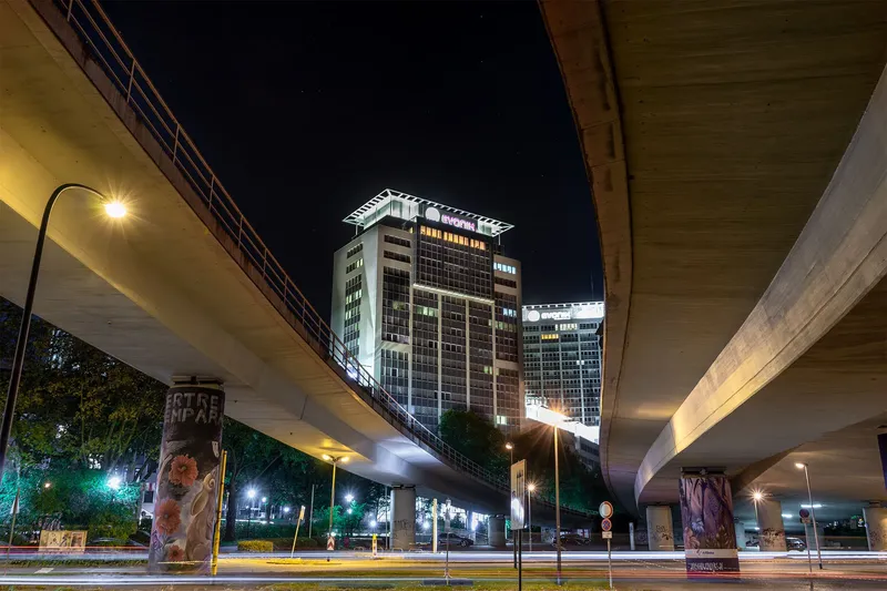 Atmosphärische Nachtaufnahme der Essener Skyline mit illuminierten Hochhäusern, fotografiert durch die urbane Architektur einer Hochstraße. Das Bild vereint moderne Stadtarchitektur, künstlerisch gestaltete Straßenpfeiler und dynamische Lichtspuren des nächtlichen Verkehrs in einer eindrucksvollen urbanen Komposition.
