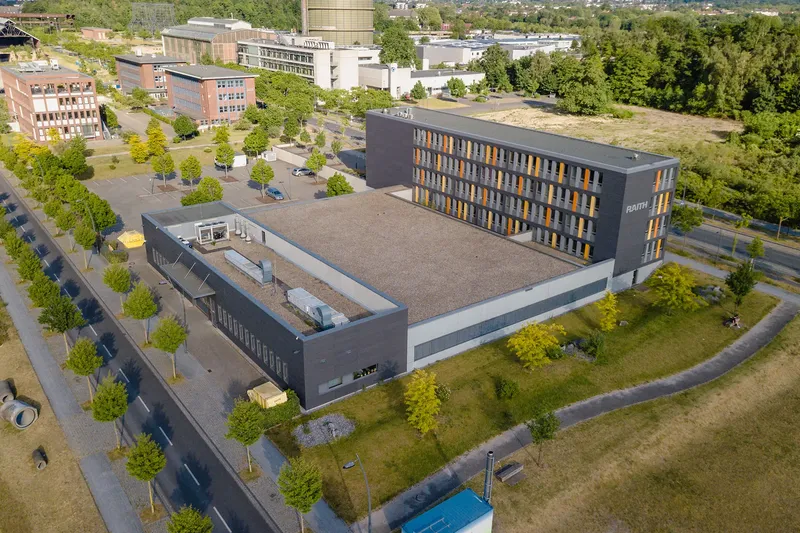 Luftaufnahme des RAITH-Technologiezentrums in Dortmund: Moderne Architektur mit charakteristischer grau-oranger Fassade, eingebettet in einen Campus-ähnlichen Technologiepark mit gepflegten Grünanlagen. Das Gebäude vereint Büro- und Forschungsflächen in zeitgemäßem Design.
