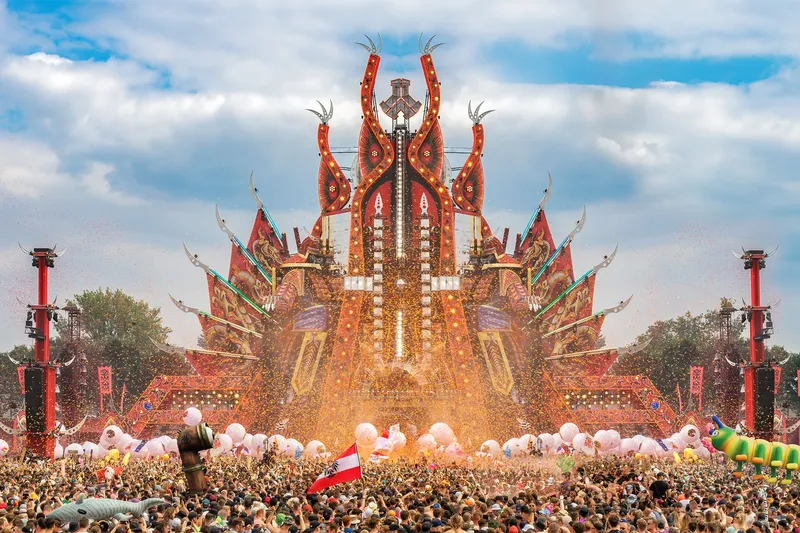 Spektakuläre Defqon.1 Mainstage während der legendären Power Hour mit imposanter roter Festival-Architektur, beeindruckenden Lichteffekten, charakteristischen aufblasbaren Figuren und einer energiegeladenen Menschenmenge unter strahlend blauem Himmel. hardstyle festival mainstage edm
