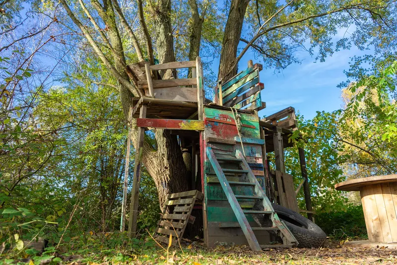 Nostalgisches Baumhaus mit bunter Holztreppe in idyllischer Waldumgebung. Handgefertigte Holzkonstruktion mit vintage Charme, umgeben von grüner Natur - perfektes Motiv für Kindheitserinnerungen und Gartengestaltung.
